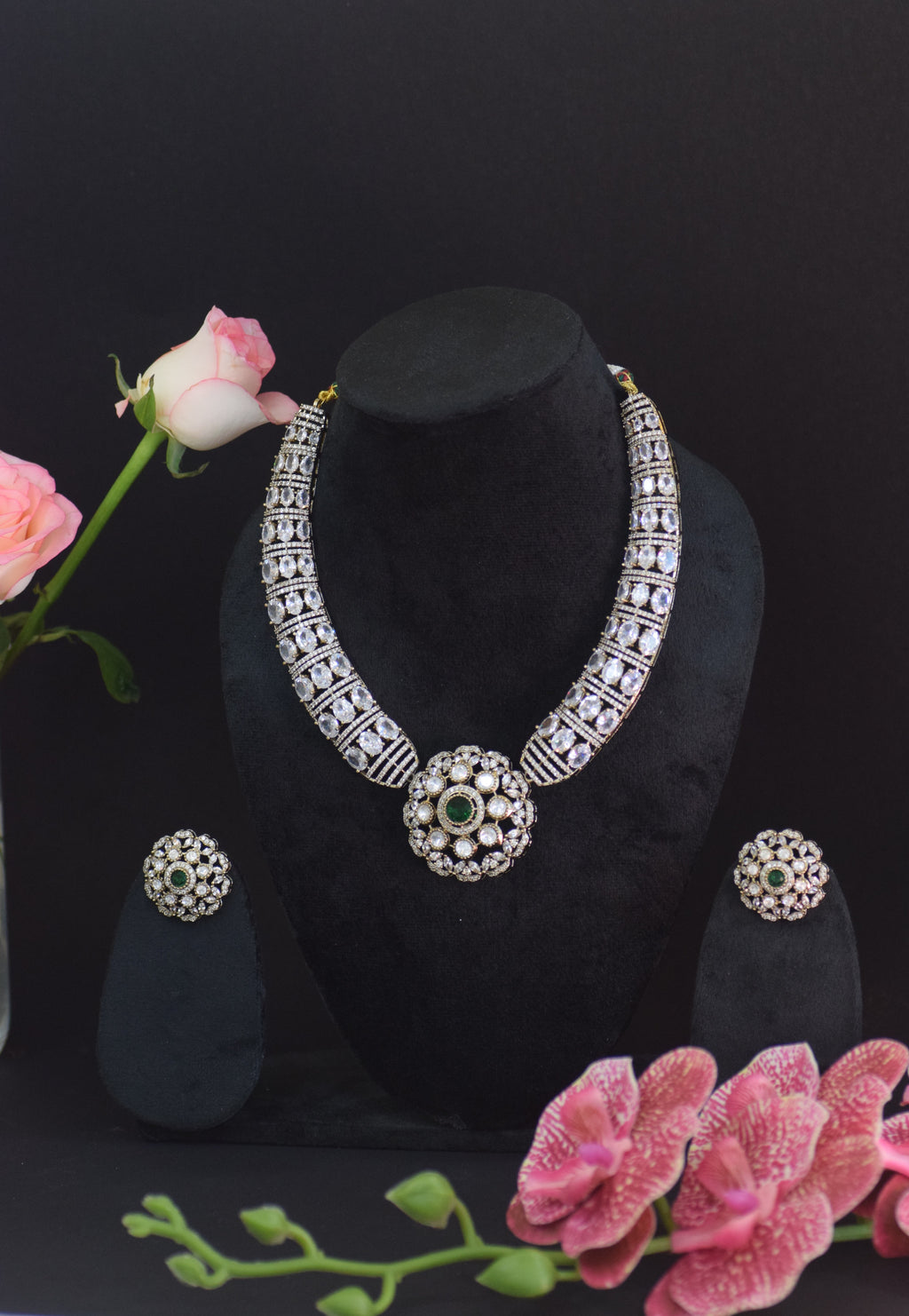 Jewelry set with necklace and earrings on a black stand, surrounded by pink flowers.