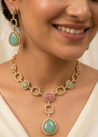 Close-up of a woman wearing a gold necklace with green and pink gemstones and matching earrings.